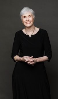 This is a portrait photo of Susan Dingle, poet-laureate of Clark County, Washington. She has short gray hair, a lovely smile, and is standing with her clasped in front of her waist, wearing an elegant black dress.